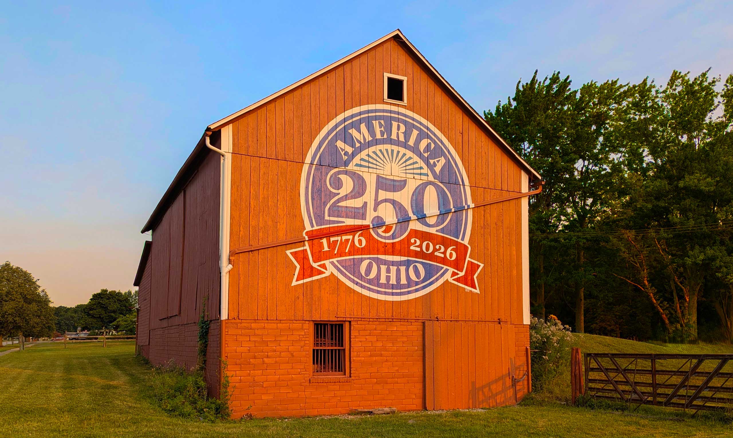 Red barn with a large blue-and-white 250th-anniversary emblem painted on its side, showing 1776–2026 and Ohio, set against a green landscape and blue sky.