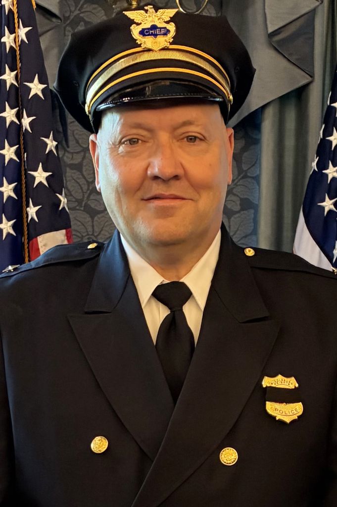 Portrait of a male police chief in formal dress uniform, wearing a peaked cap with 'CHIEF' badge, standing in front of American flags.